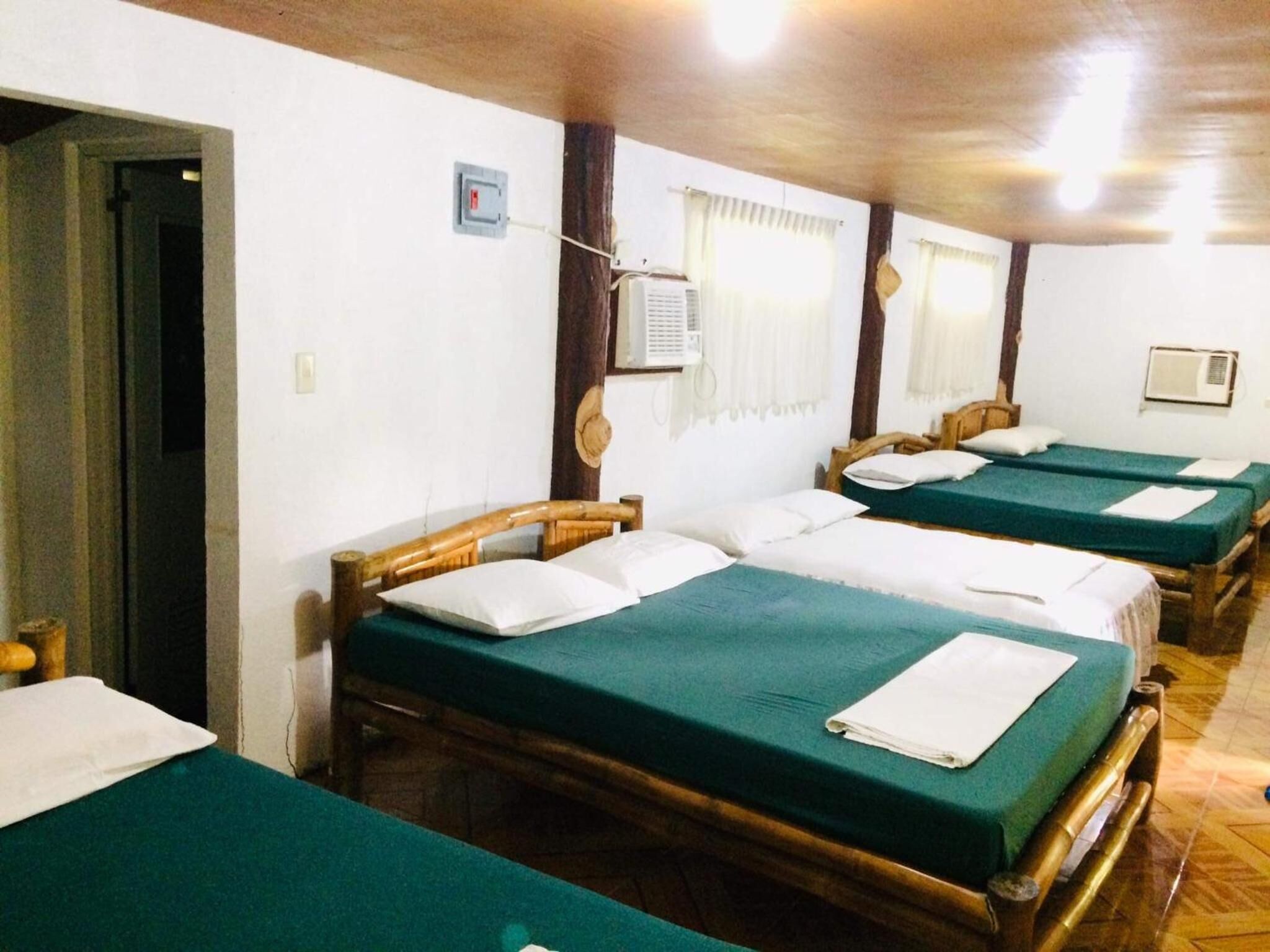 Quarto com quatro camas de solteiro, cada uma com colcha verde e almofadas brancas. Possui duas unidades de ar condicionado fixadas na parede, uma em cada parede.