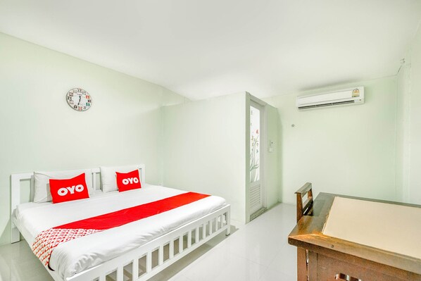 A hotel room with a bed, a desk, an air conditioner, and a wall clock.