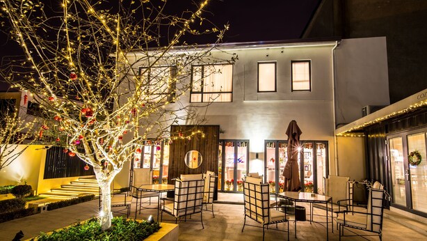 An outdoor seating area with tables and chairs, a decorative tree with lights, and a building with large windows and a door.