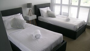 Dos camas individuales con sábanas y almohadas blancas, una mesita de noche con lámpara y una ventana con persianas.