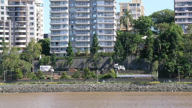 Un edificio residencial de varios pisos con balcones, un terraplén rocoso y una masa de agua al frente.