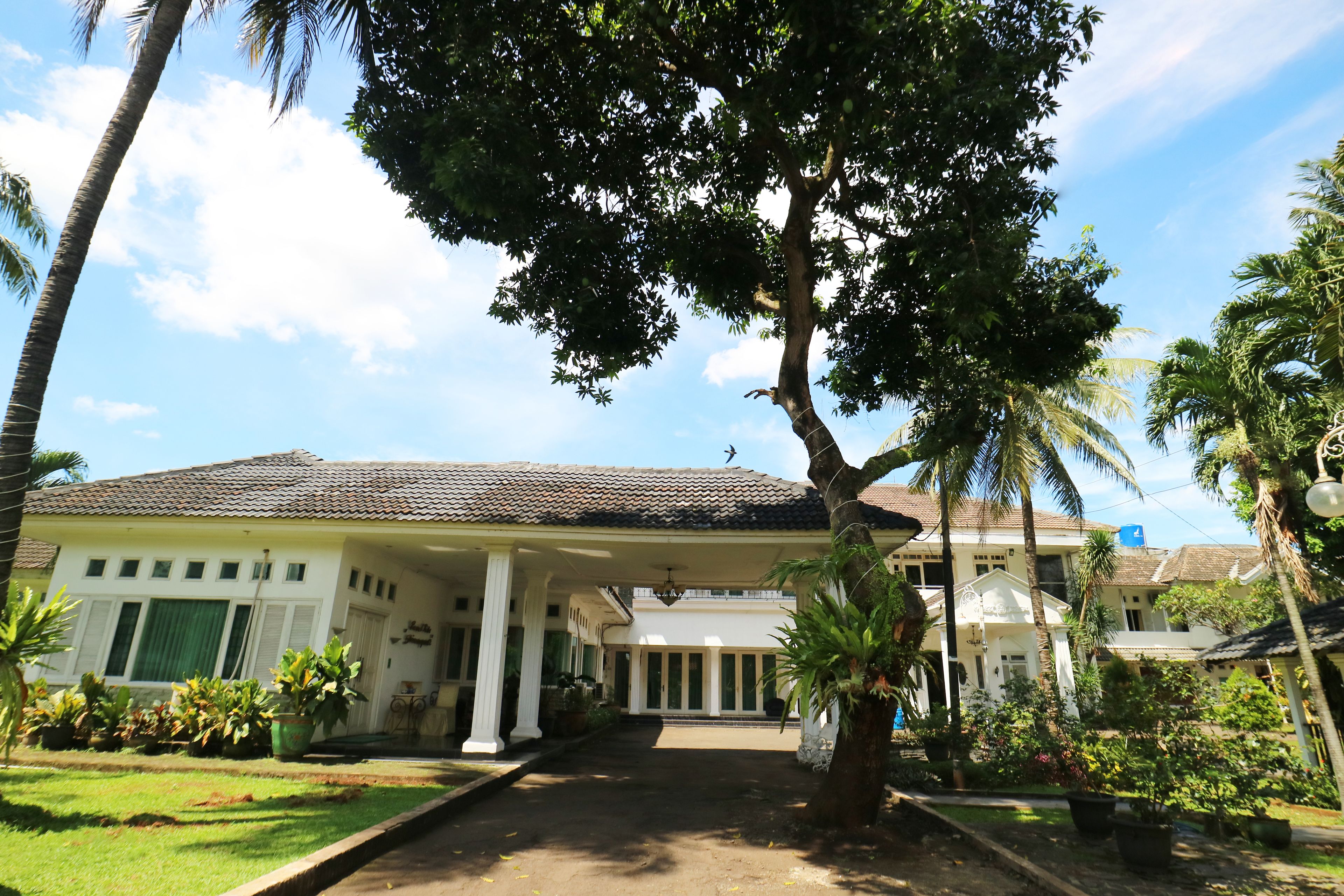 A colonial-style building with a tiled roof, a covered entrance with columns, and a well-maintained garden with palm trees and potted plants.