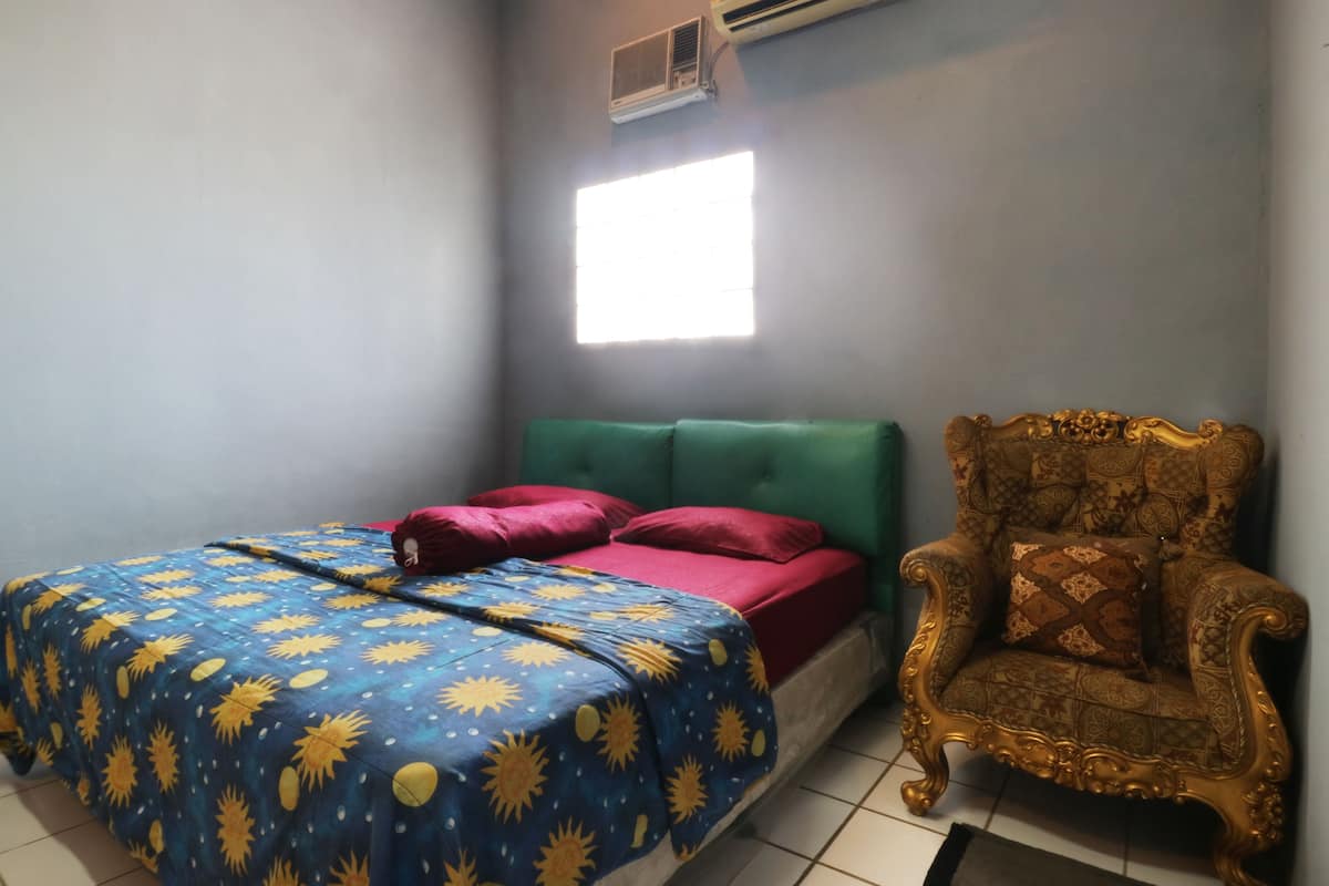 Ein Zimmer mit einem Bett, einem grünen gepolsterten Kopfteil, einem gemusterten Sessel und einem Fenster mit Tageslicht.