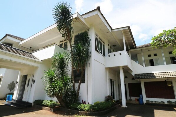 A white multi-story residential building with a flat roof, a balcony, and a small garden area with palm trees.