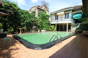 A swimming pool with a brick deck, surrounded by greenery and a building with a balcony.