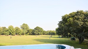 Piscina rodeada por un área verde con árboles y gente al fondo.