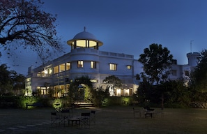 A large, white building with a central dome and multiple windows, surrounded by a well-maintained garden with benches and chairs.
