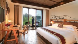 Habitación de hotel con una cama grande, un escritorio de madera con silla, un balcón con vistas a zonas verdes y un ventanal amplio.