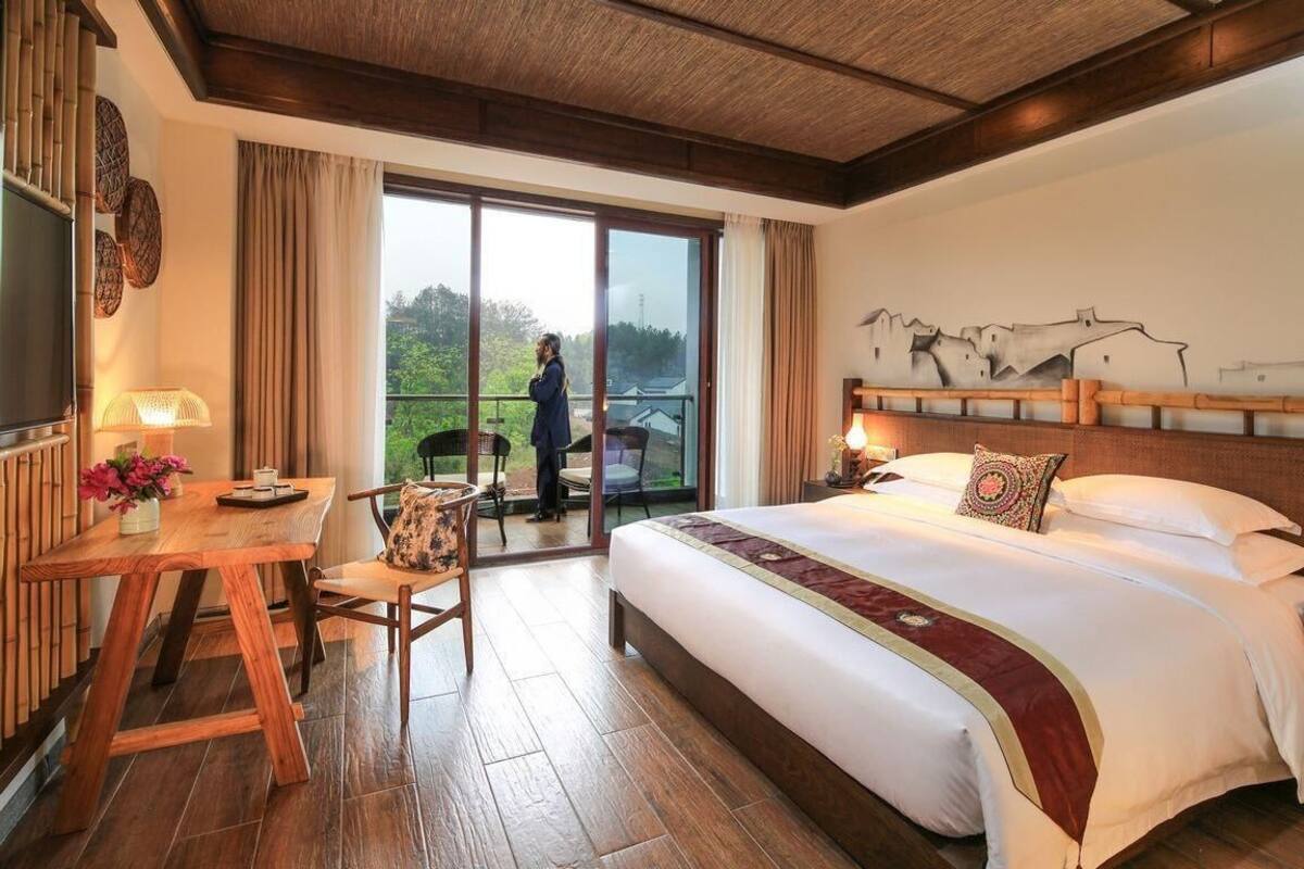 Habitación de hotel con una cama grande, un escritorio de madera con silla, un balcón con vistas a zonas verdes y un ventanal amplio.