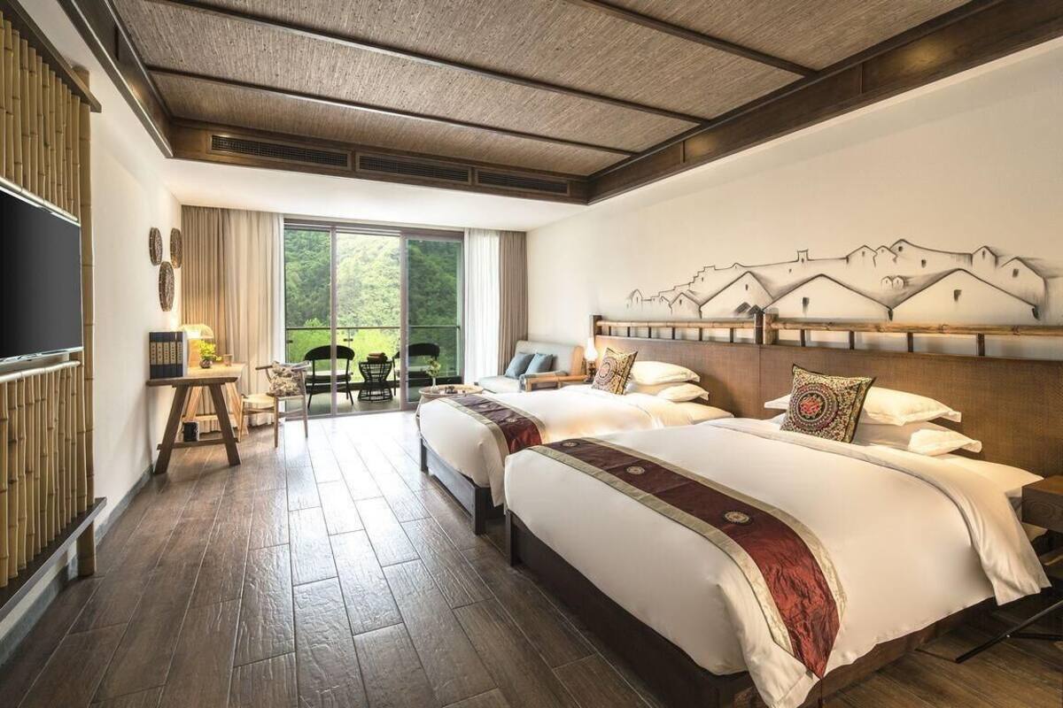 Habitación de hotel con dos camas, un ventanal con vistas a la vegetación, un televisor de pantalla plana y una obra de arte colgada en la pared que representa montañas.