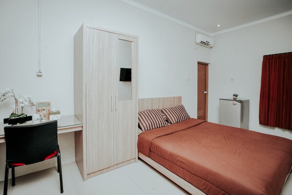 A bedroom with a bed, a desk, a chair, a wardrobe, a mirror, and a small refrigerator.