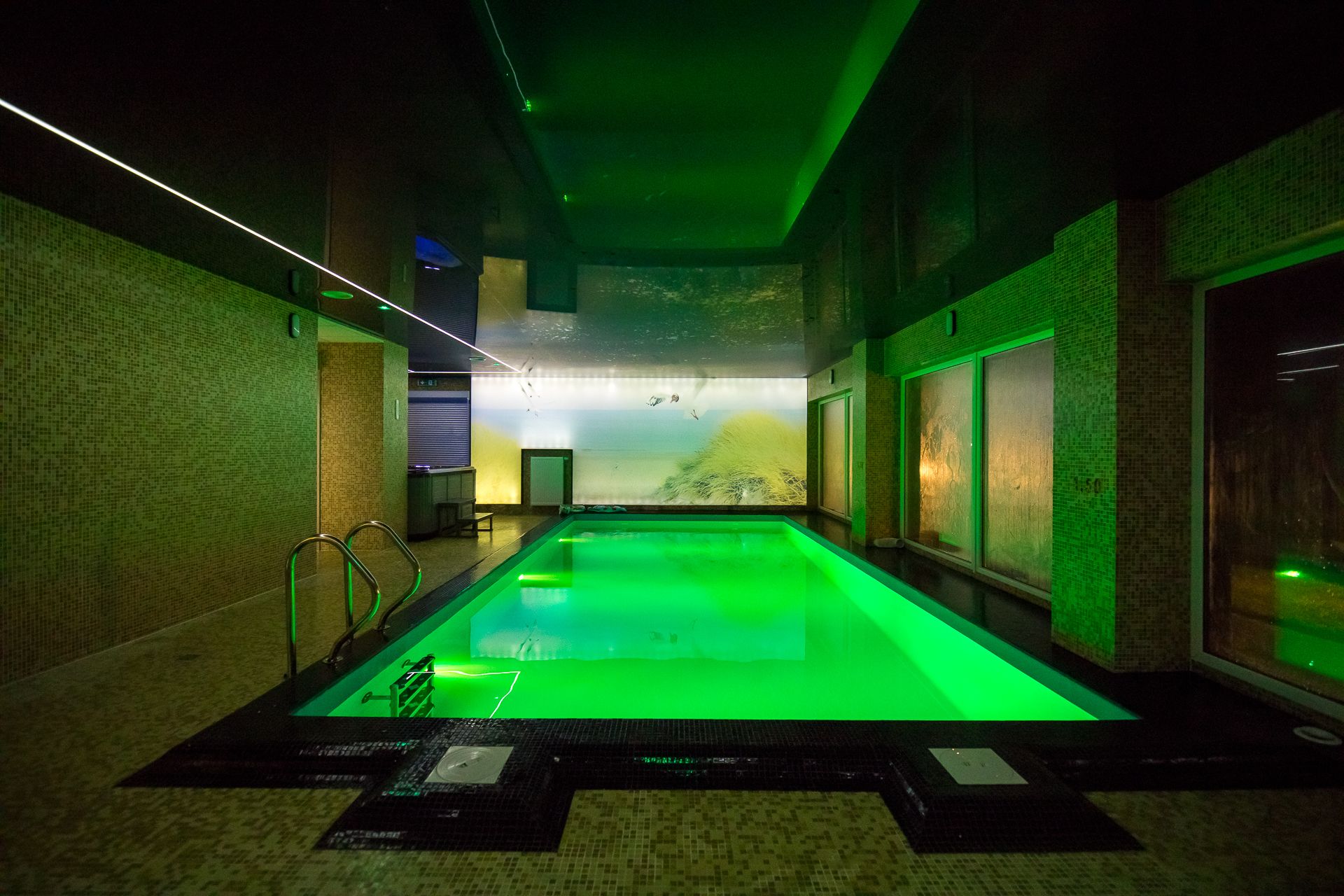 A swimming pool with a view of a beach through a glass wall, surrounded by tiled walls and a green-lit ceiling.