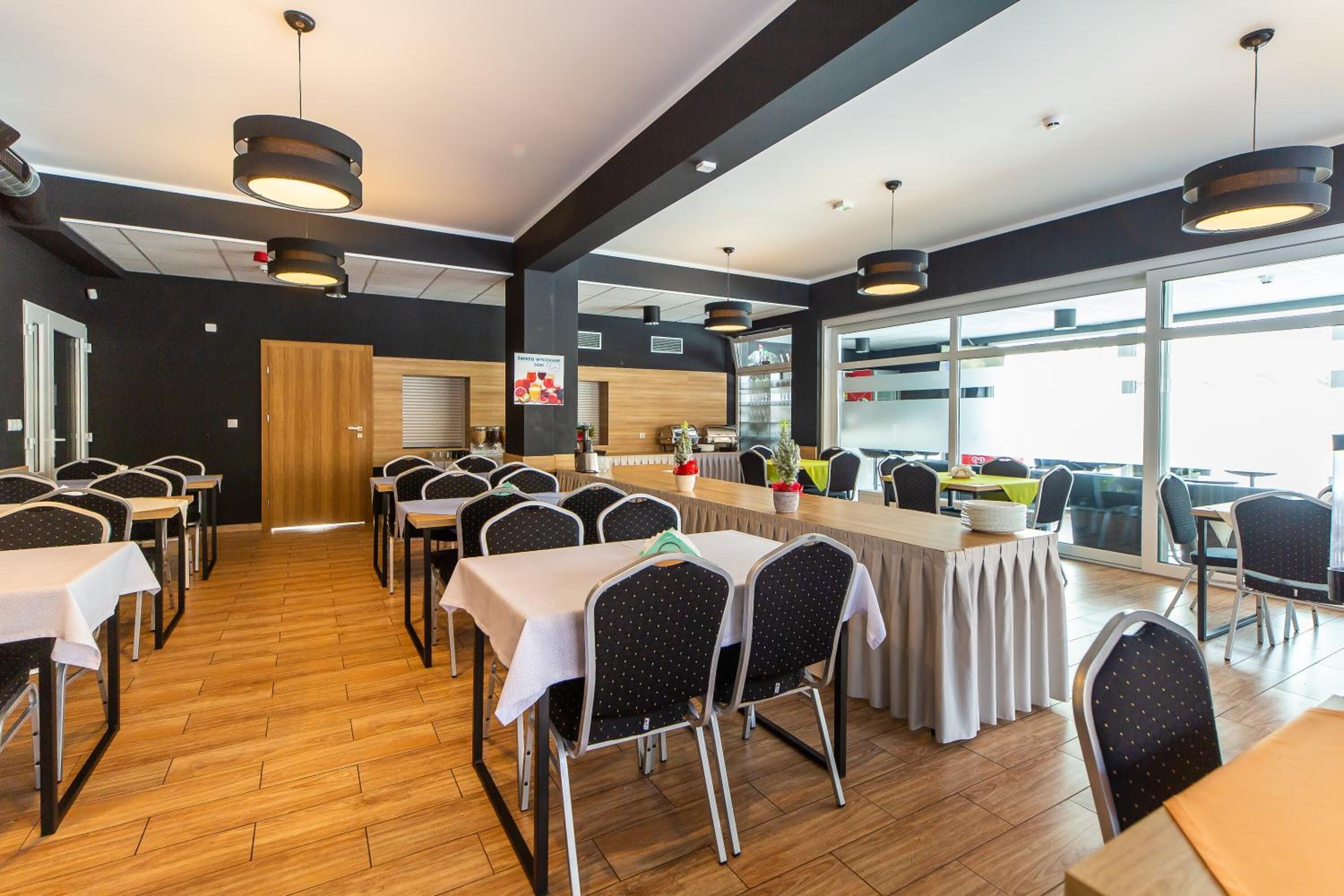 A conference room with multiple tables and chairs, large windows, and a kitchen area in the background.