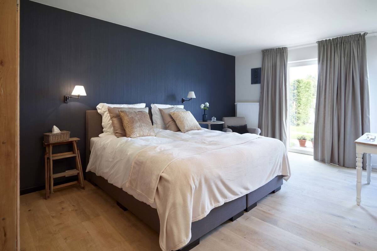 Quarto com cama grande, mesas de cabeceira, uma cadeira e vista para o exterior.