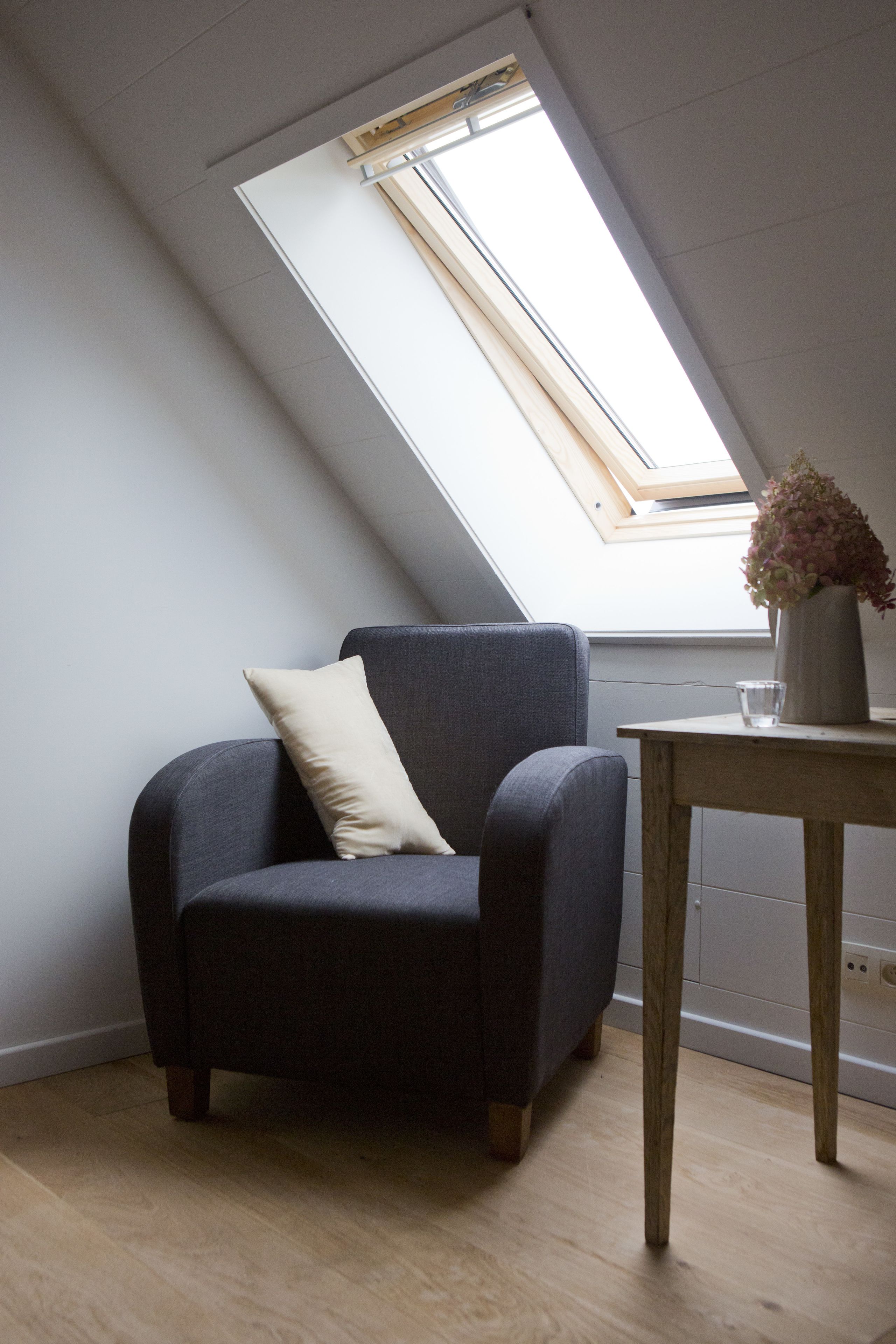 A room with a skylight, a dark grey armchair, a wooden table, and a vase with flowers.