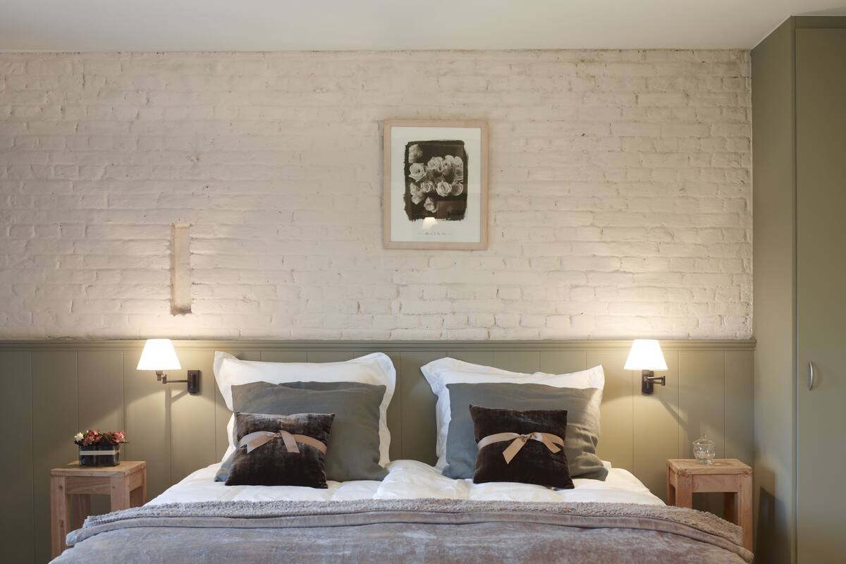 Uma cama de casal com cabeceira estofada cinza, duas mesas de cabeceira com luminárias, uma obra de arte emoldurada acima da cama e uma mesinha de cabeceira de madeira com um vaso de flores.