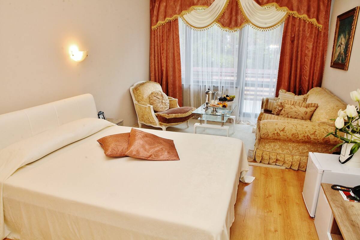 Habitación de hotel con una cama grande, un sofá, una silla, una mesita con una bandeja de fruta y una ventana con cortinas.