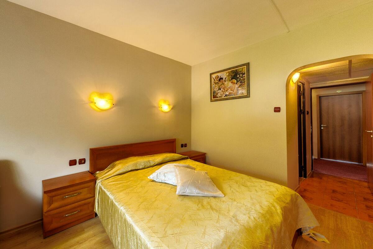 Una habitación de hotel con cama, mesitas de noche, un cuadro en la pared y una puerta de madera.