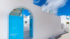 Edifici blanc amb una porta i un arc blaus, que condueix a una zona d'estar a l'aire lliure amb cadires taronja.
