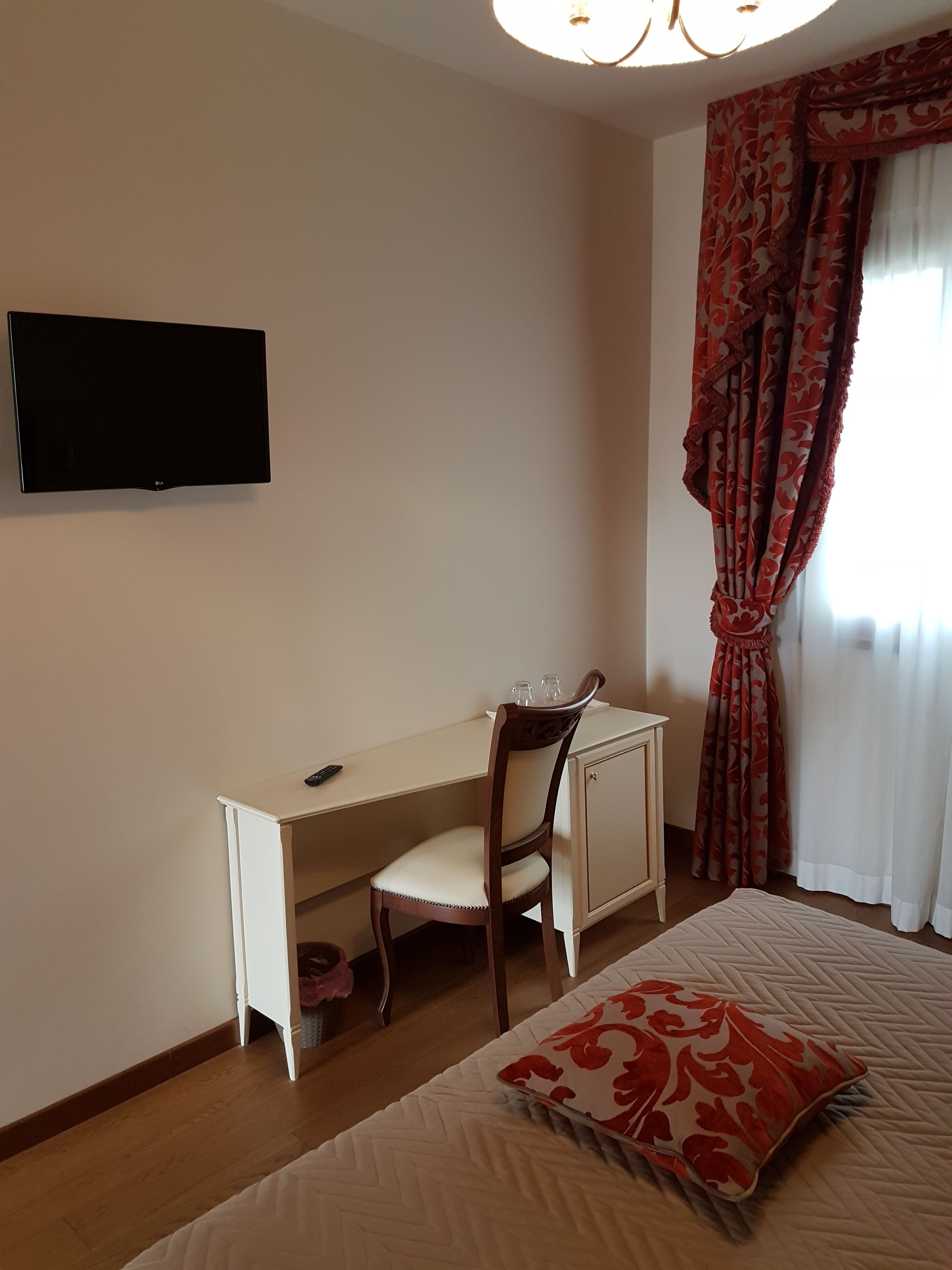 Una camera da letto con un letto, una scrivania con una sedia, una televisione a parete e una finestra con tende.