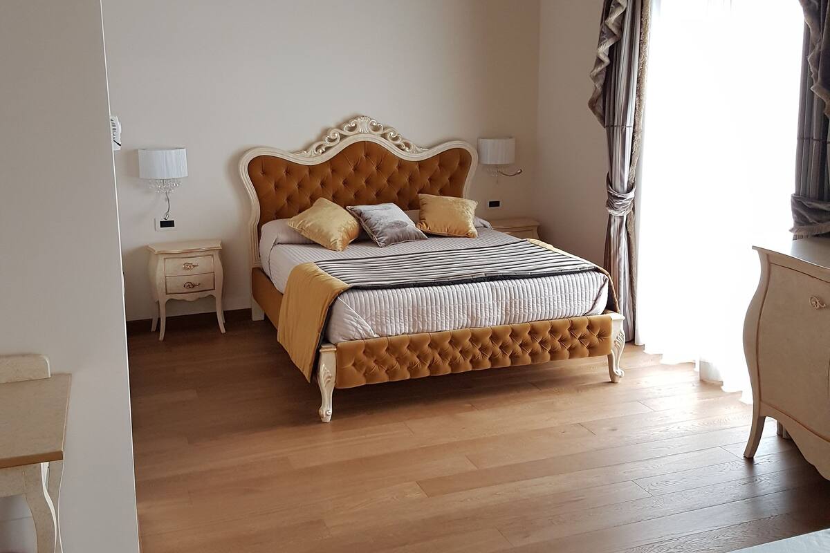Un dormitorio con piso de madera, una cama con cabecera capitonada, dos mesitas de noche con lámparas, un tocador y una ventana con cortinas.