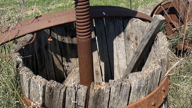 Estrutura metálica antiga e enferrujada com uma grande roda e uma barra vertical, cercada por um campo gramado e uma cerca.