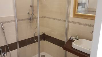 Box doccia in vetro con soffione a mano, portasci a parete e lavabo bianco con specchio sopra.