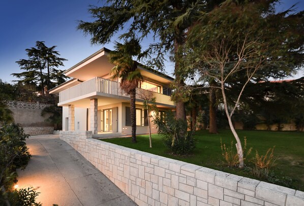Une maison à deux étages dotée d’un balcon, entourée d’arbres et d’une pelouse bien entretenue.