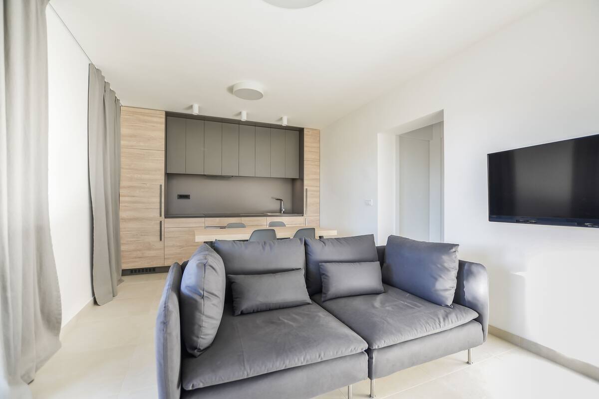 A modern living room with a grey sofa, a flat-screen TV mounted on the wall, and a wooden kitchen area in the background.