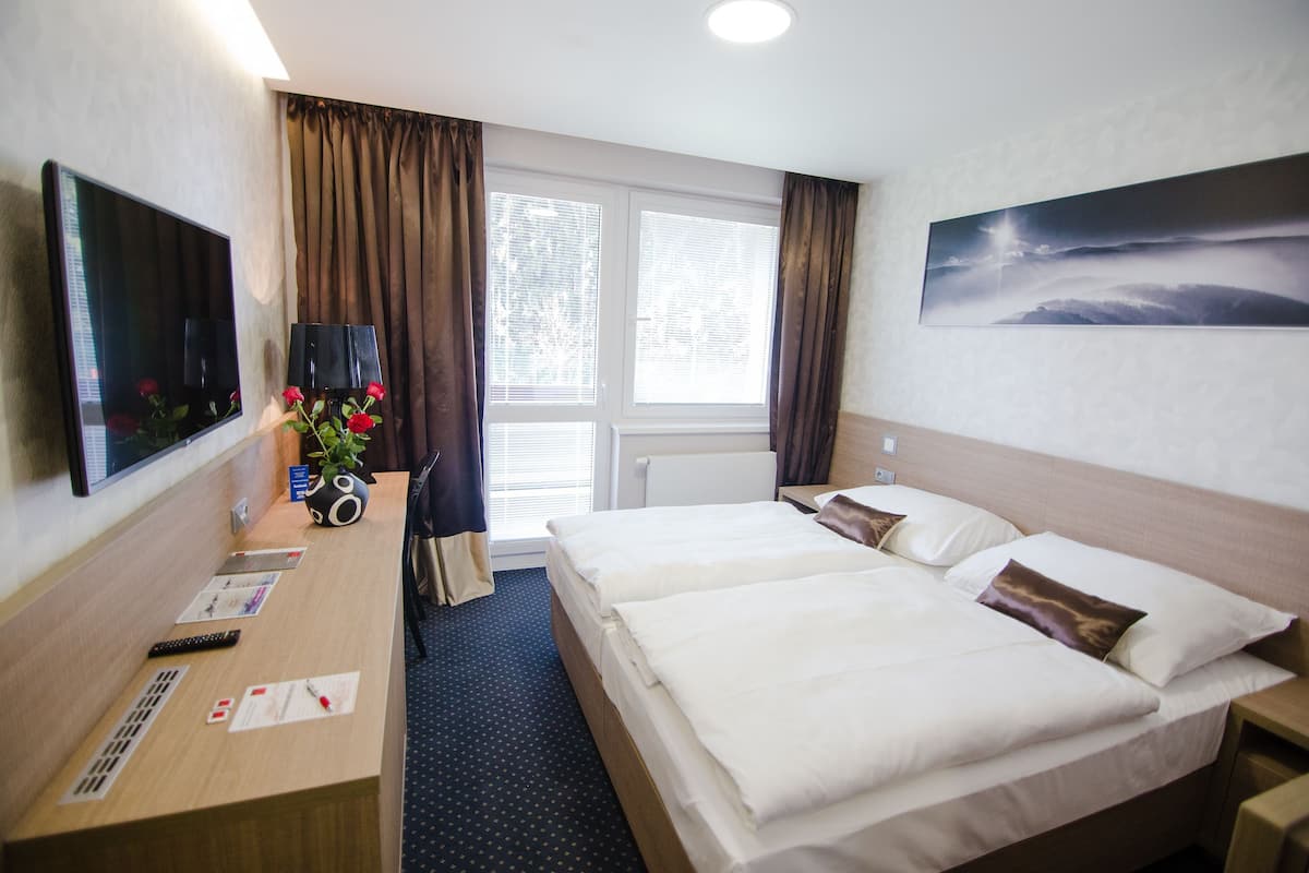 A hotel room with a bed, a desk with a TV, a vase with flowers, and a window with curtains.