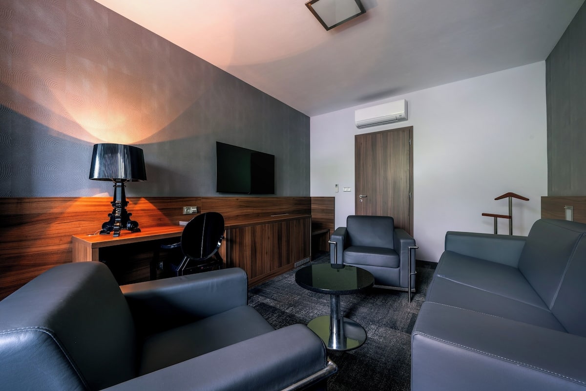 A modern hotel room with a sofa, a small table, a lamp, a TV, and a wooden panel wall.
