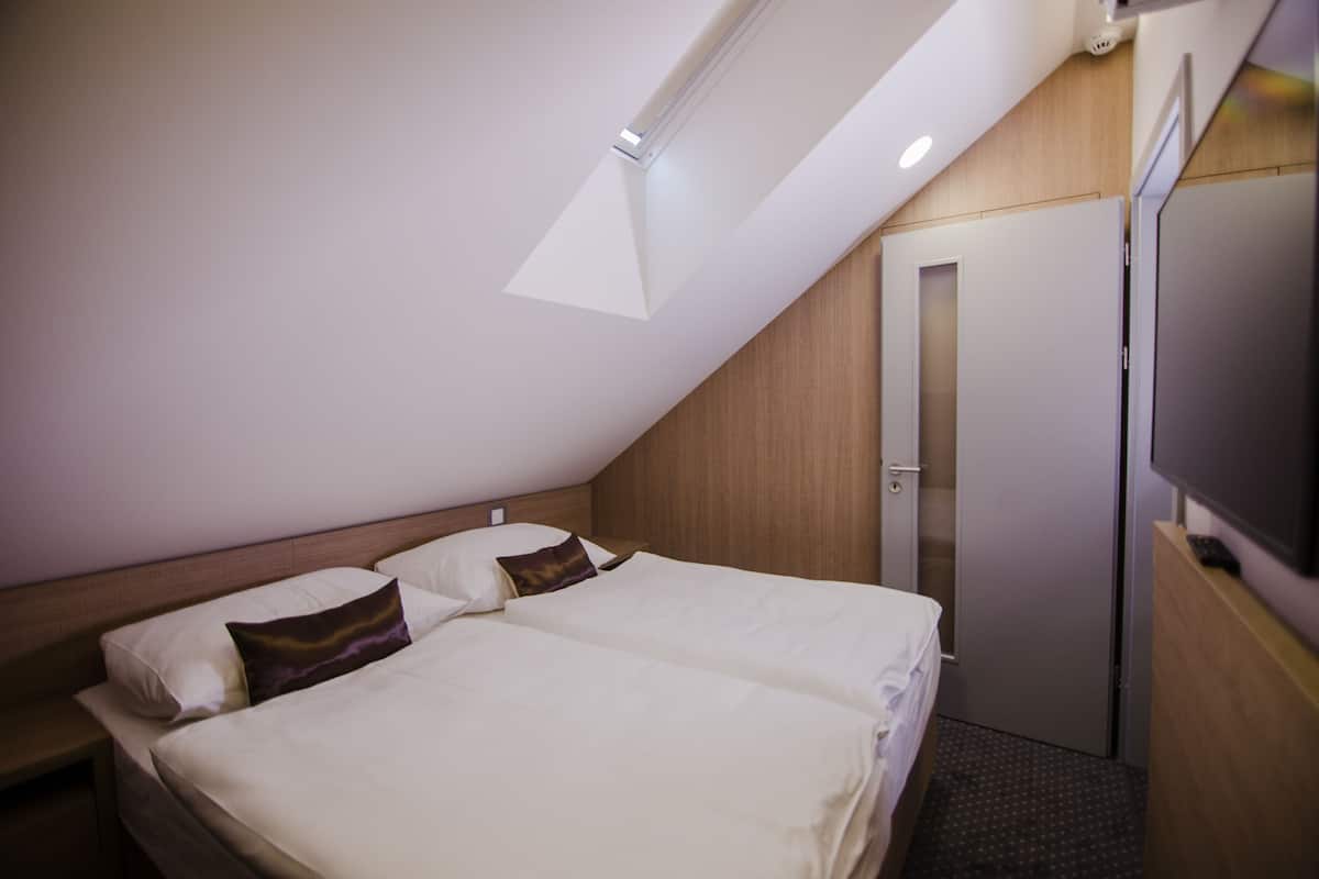 A hotel room with a sloped ceiling, a bed with white bedding and two pillows, a wooden nightstand, a television on a wooden stand, and a door leading to another room.