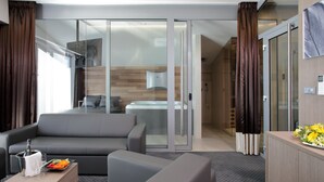 A modern hotel room with a grey sofa, a small table with a bottle and glasses, a tray with fruit, and a view of a kitchen and bathroom through a glass door. - Hotel Jezerka (Sec)