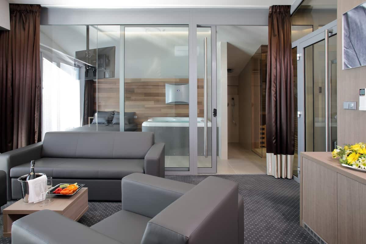 A modern hotel room with a grey sofa, a small table with a bottle and glasses, a tray with fruit, and a view of a kitchen and bathroom through a glass door.
