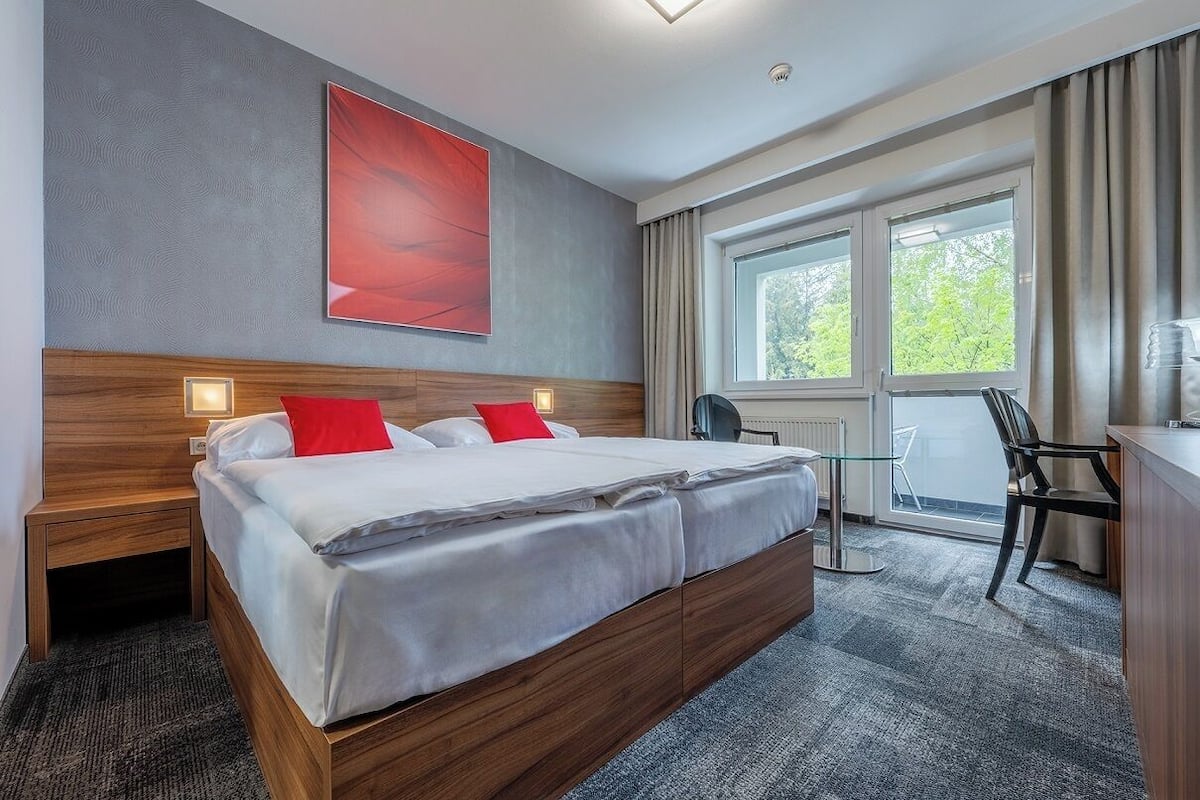 A modern hotel room with a large bed, a desk, and a window with a view of trees.
