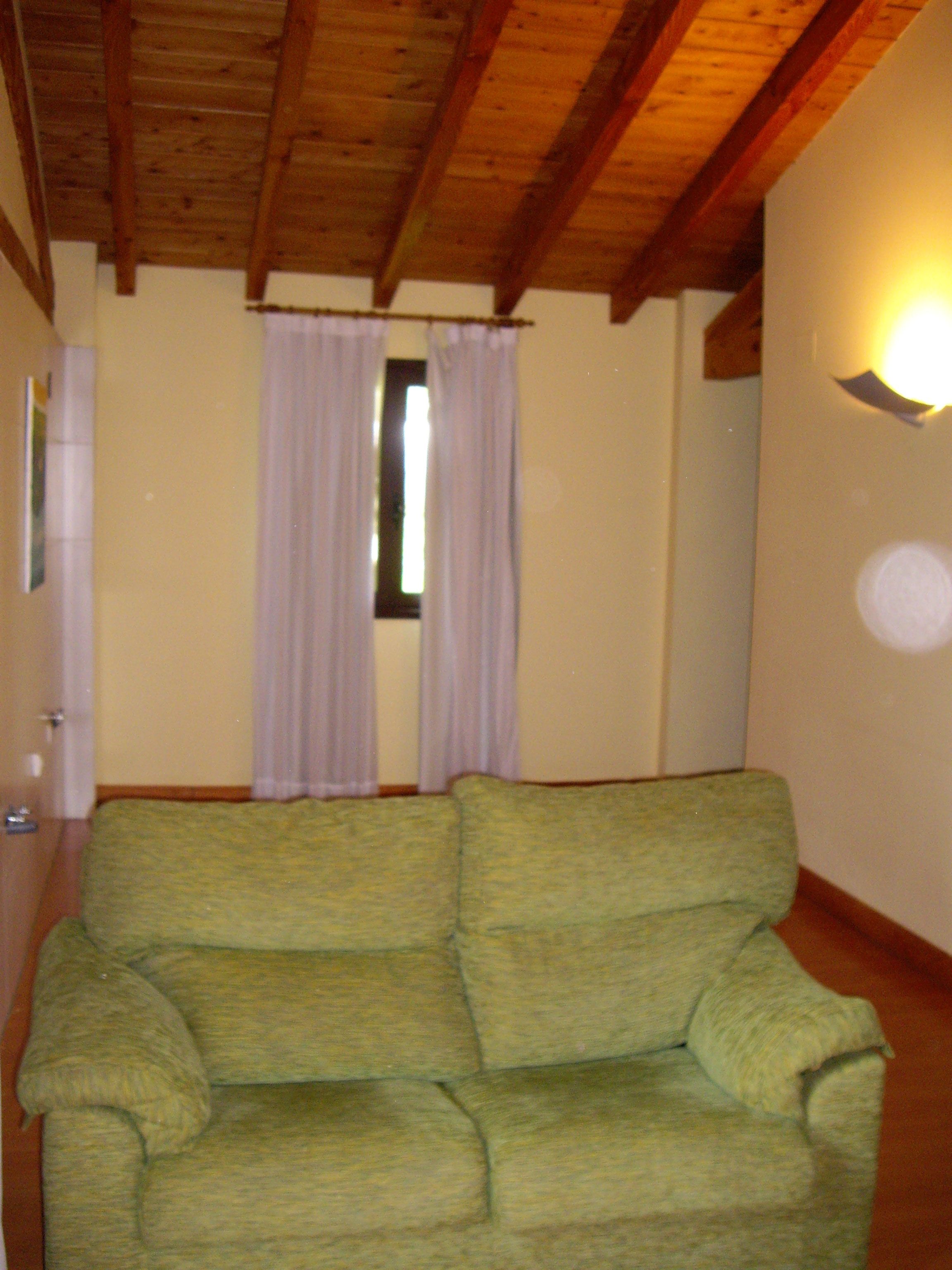 Una habitación con techo de madera, un sofá verde, una ventana con cortinas blancas y una lámpara fijada a la pared.