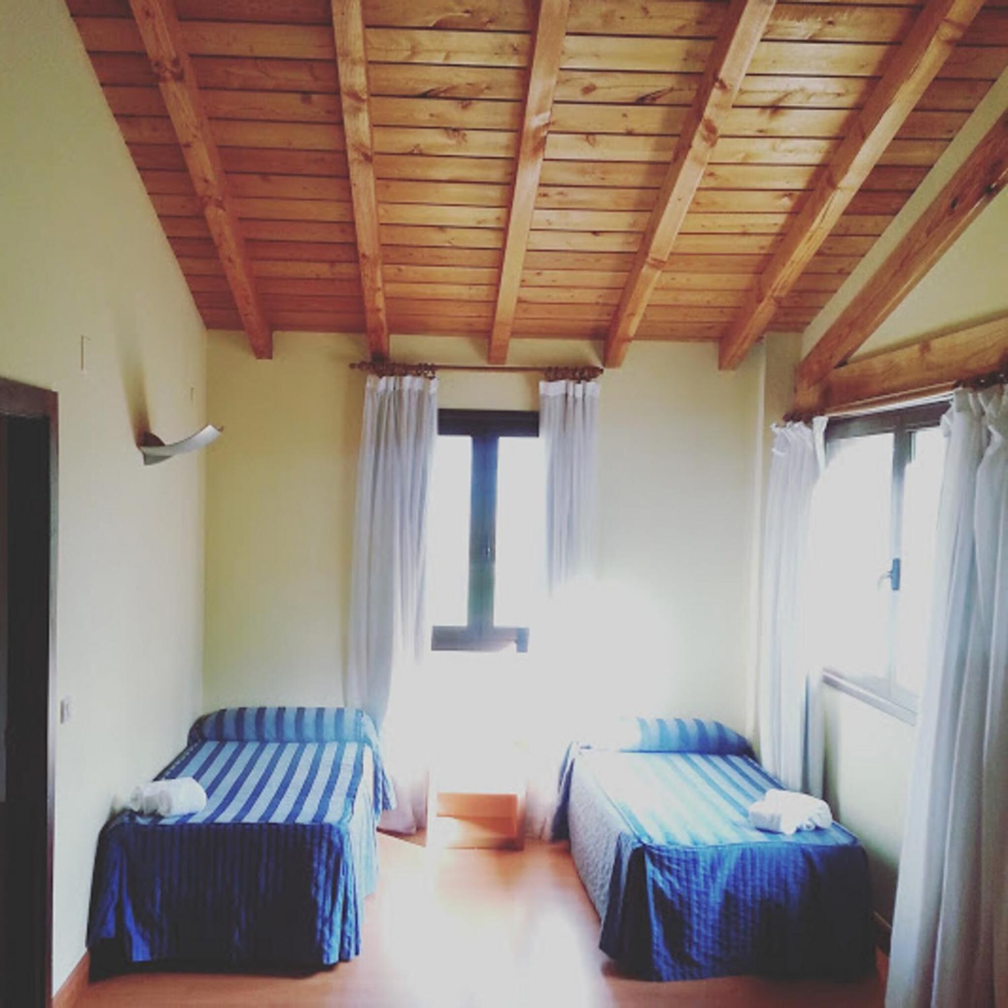 Une chambre avec deux lits, des poutres apparentes en bois au plafond, une fenêtre avec des rideaux et une porte.