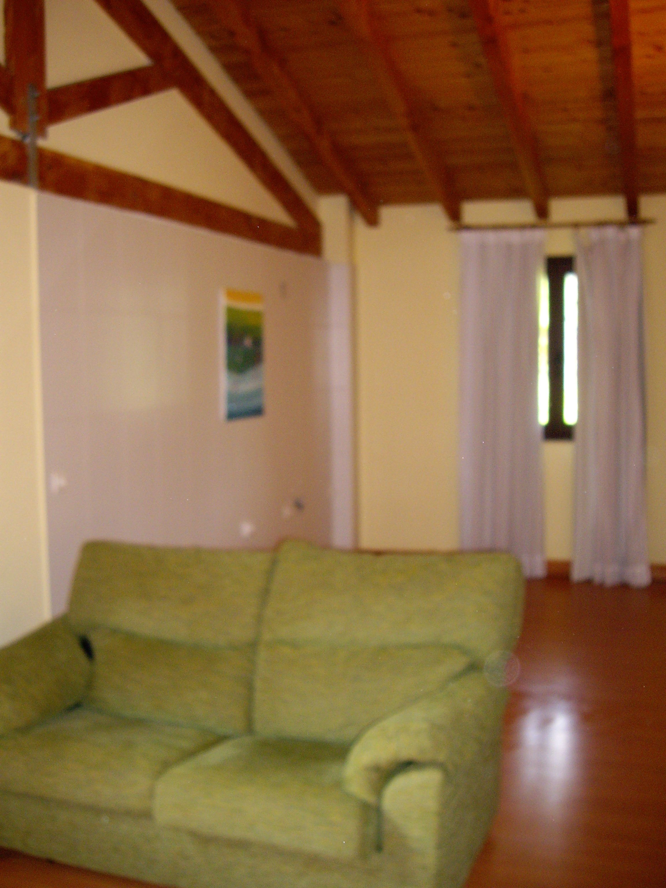 Um sofá verde num quarto com vigas de madeira no teto, uma janela com cortinas e uma parede com um cartaz.