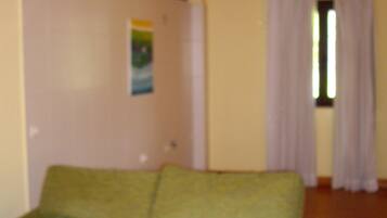 Um sofá verde num quarto com vigas de madeira no teto, uma janela com cortinas e uma parede com um cartaz.