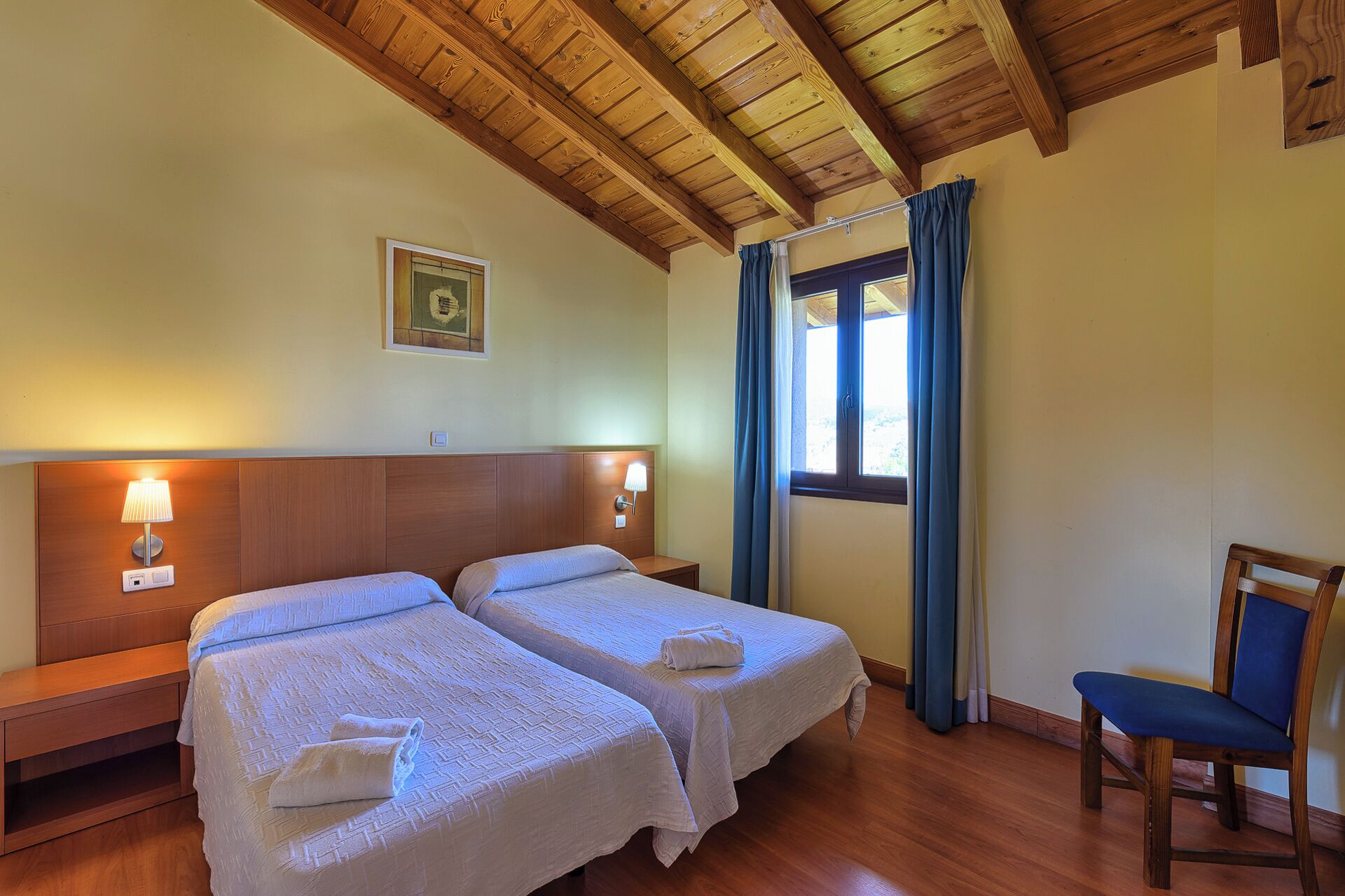 Une chambre d’hôtel avec deux lits, des têtes de lit en bois, une table de chevet avec une lampe, une chaise et une fenêtre avec des rideaux.