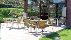 An outdoor patio with wicker chairs and tables, a brick building with large windows, and a view of a hillside. - Hotel Ibaigune (Murueta)