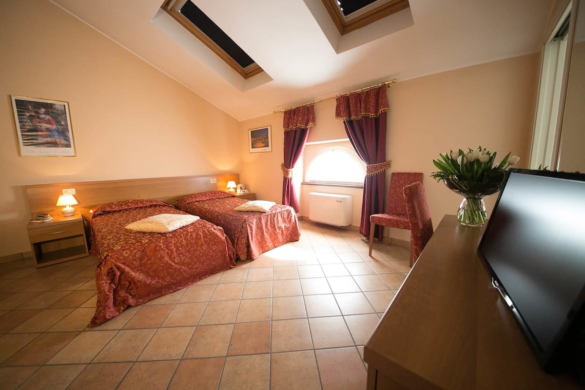 A hotel room with two beds, a desk, a chair, a TV, and a window with curtains.