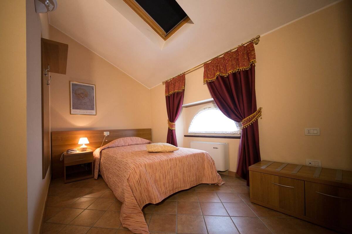 A bedroom with a bed, bedside table, lamp, window with curtains, radiator, and a small cabinet.