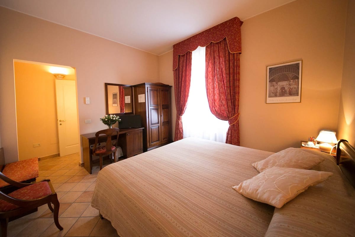 A bedroom with a large bed, a desk, a chair, a wardrobe, and a nightstand with a lamp.