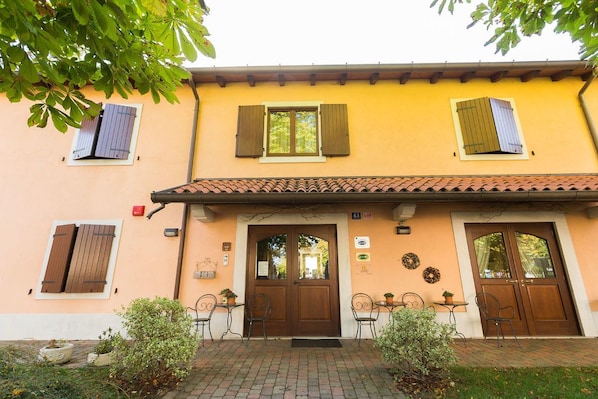 A building with a yellow facade, a tiled roof, and a central entrance with double doors. There are two windows with shutters, and a small outdoor seating area with tables and chairs.