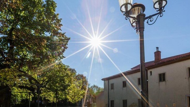 Un espacio exterior soleado con una fuente reflectante, un farol y un edificio al fondo.