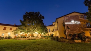Un hôtel à la façade en pierre, une enseigne indiquant « DÉPENDANCE Lipa », et un espace extérieur bien éclairé, agrémenté d’arbres et de jeux de lumière.