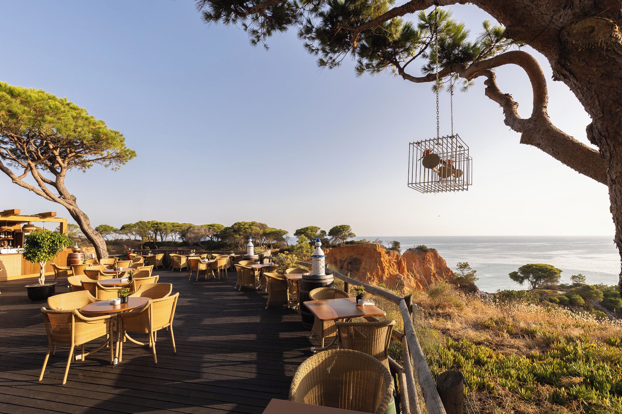 Un área de comedor al aire libre con mobiliario de mimbre, vista al océano y un árbol con una jaula colgante.