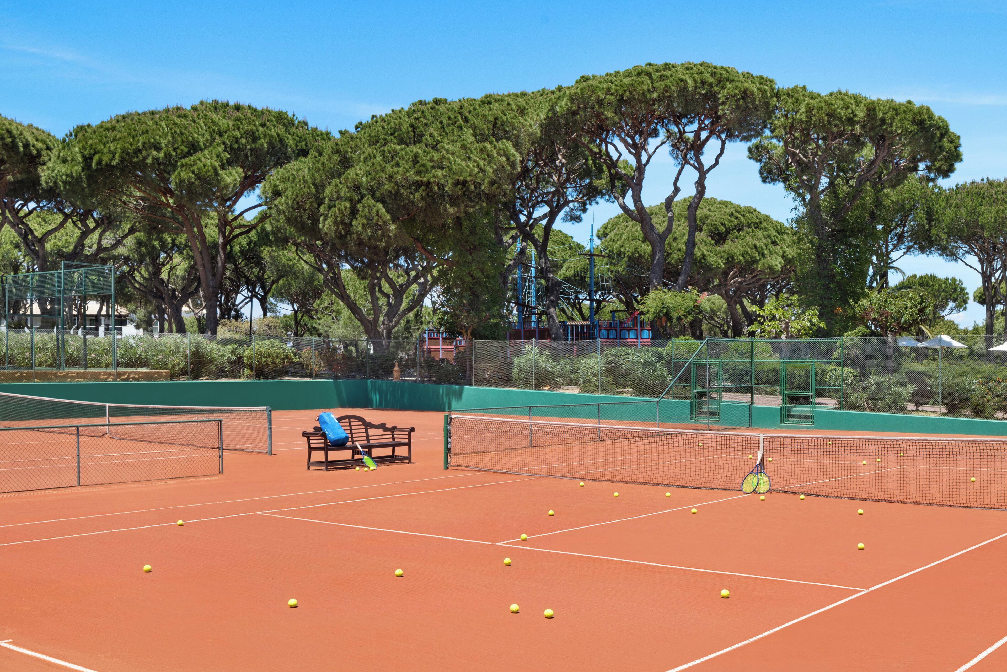 A tennis court with a bench, trees, and a fence.