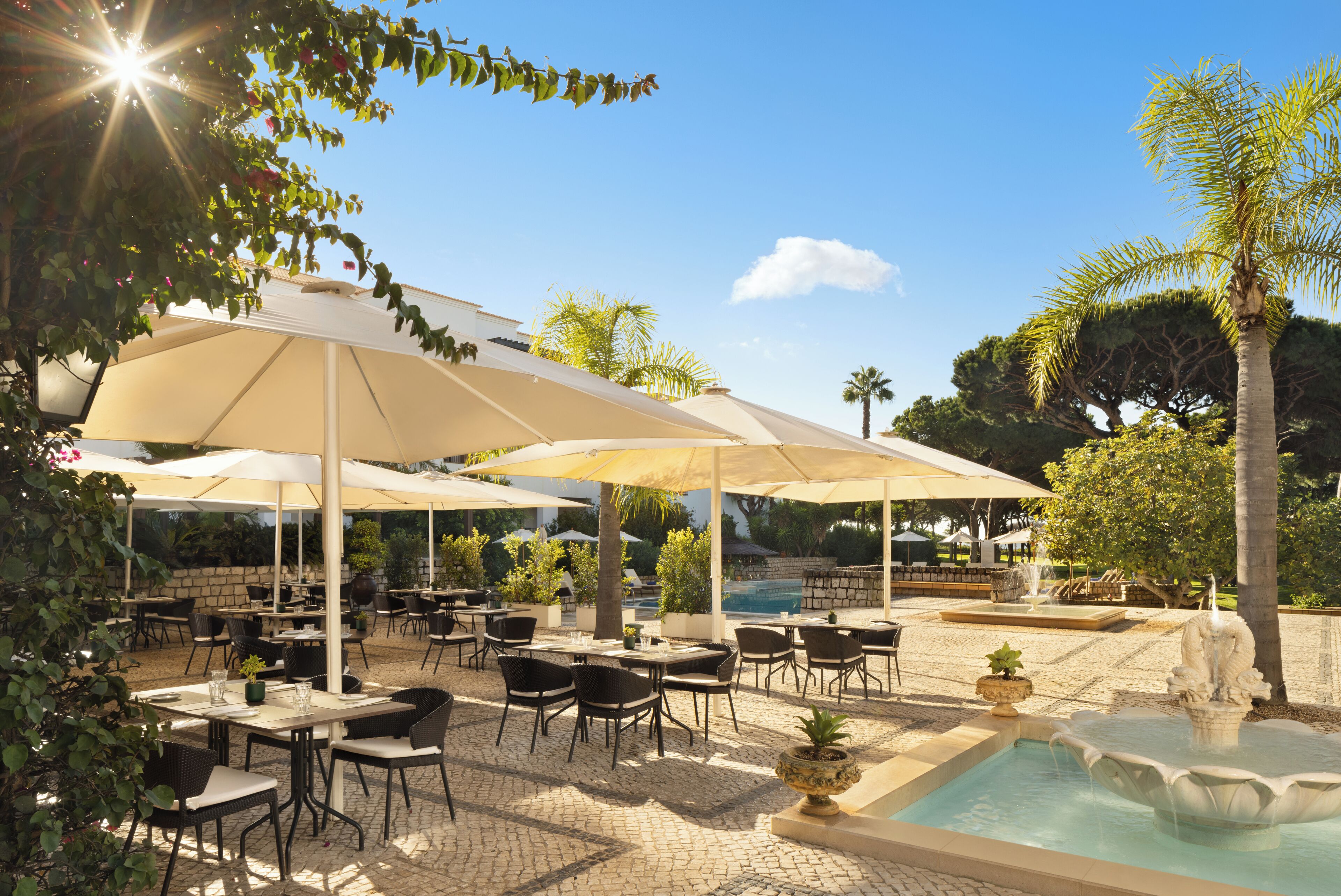 An outdoor dining area with tables and chairs, umbrellas, and a small pool with a fountain.